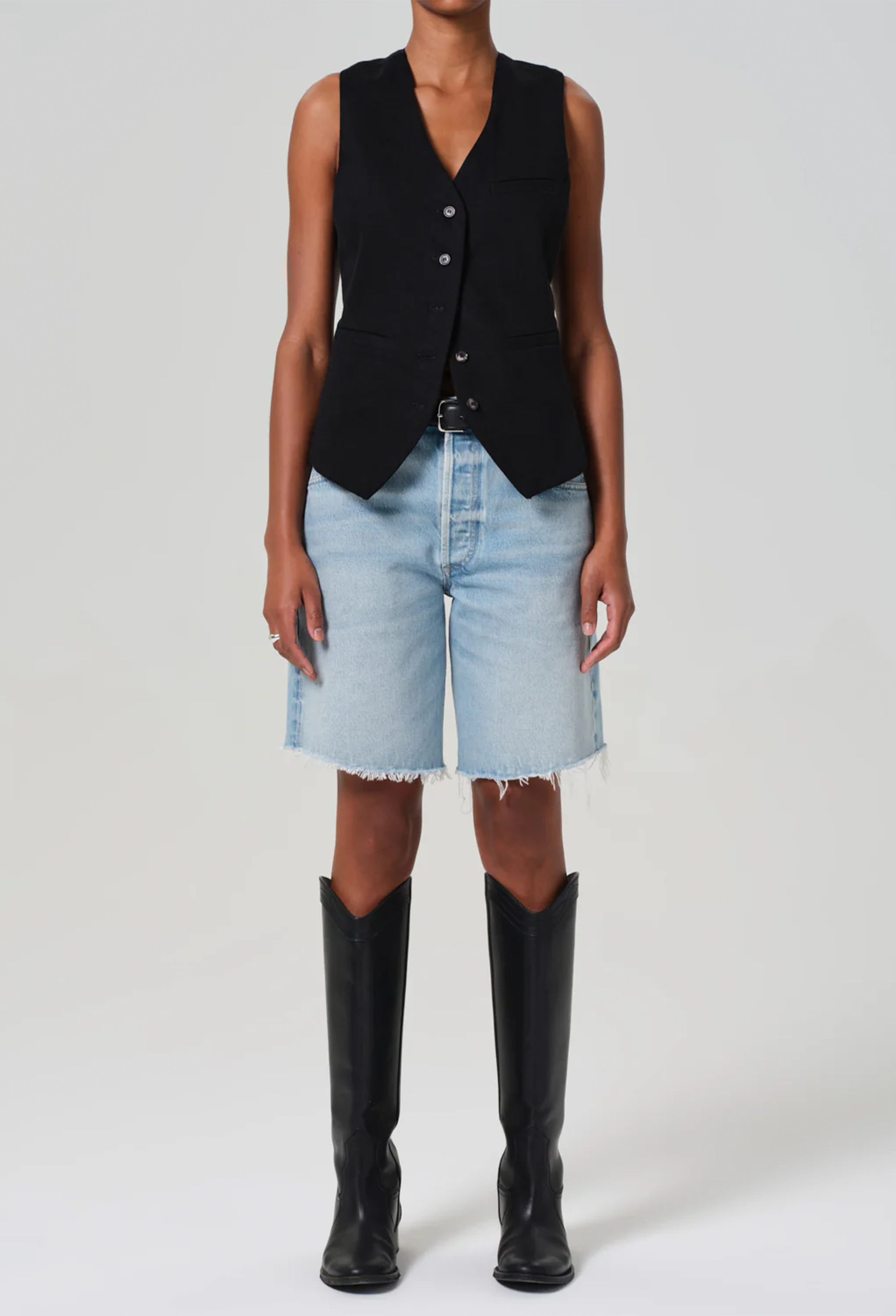 a model wearing a black vest and long light wash denim shorts with a frayed hem. white background.