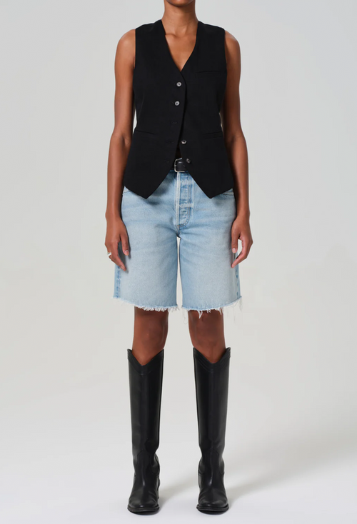 a model wearing a black vest and long light wash denim shorts with a frayed hem. white background.
