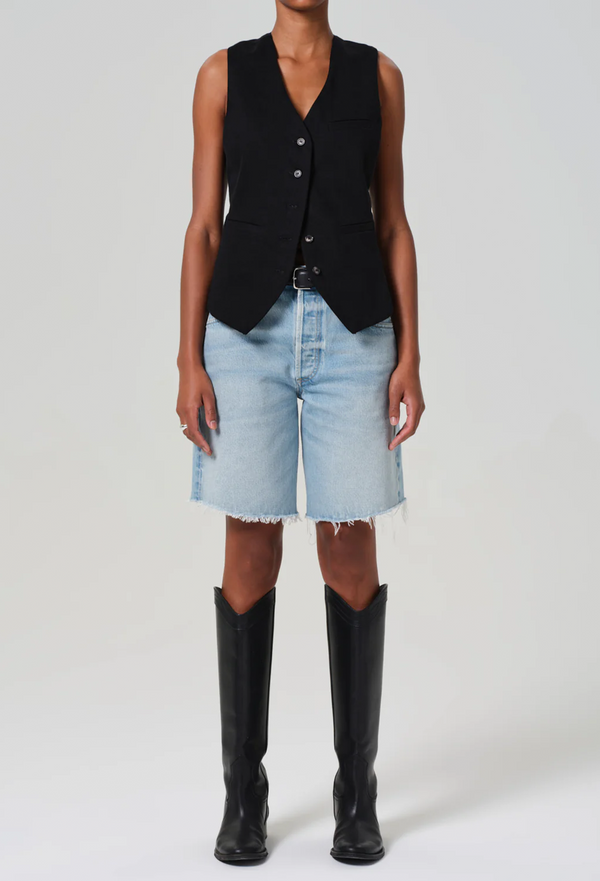 a model wearing a black vest and long light wash denim shorts with a frayed hem. white background.