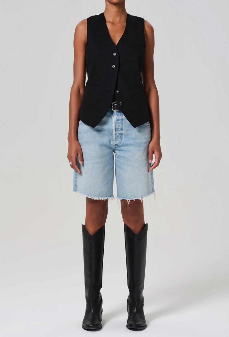 a model wearing a black vest and long light wash denim shorts with a frayed hem. white background.