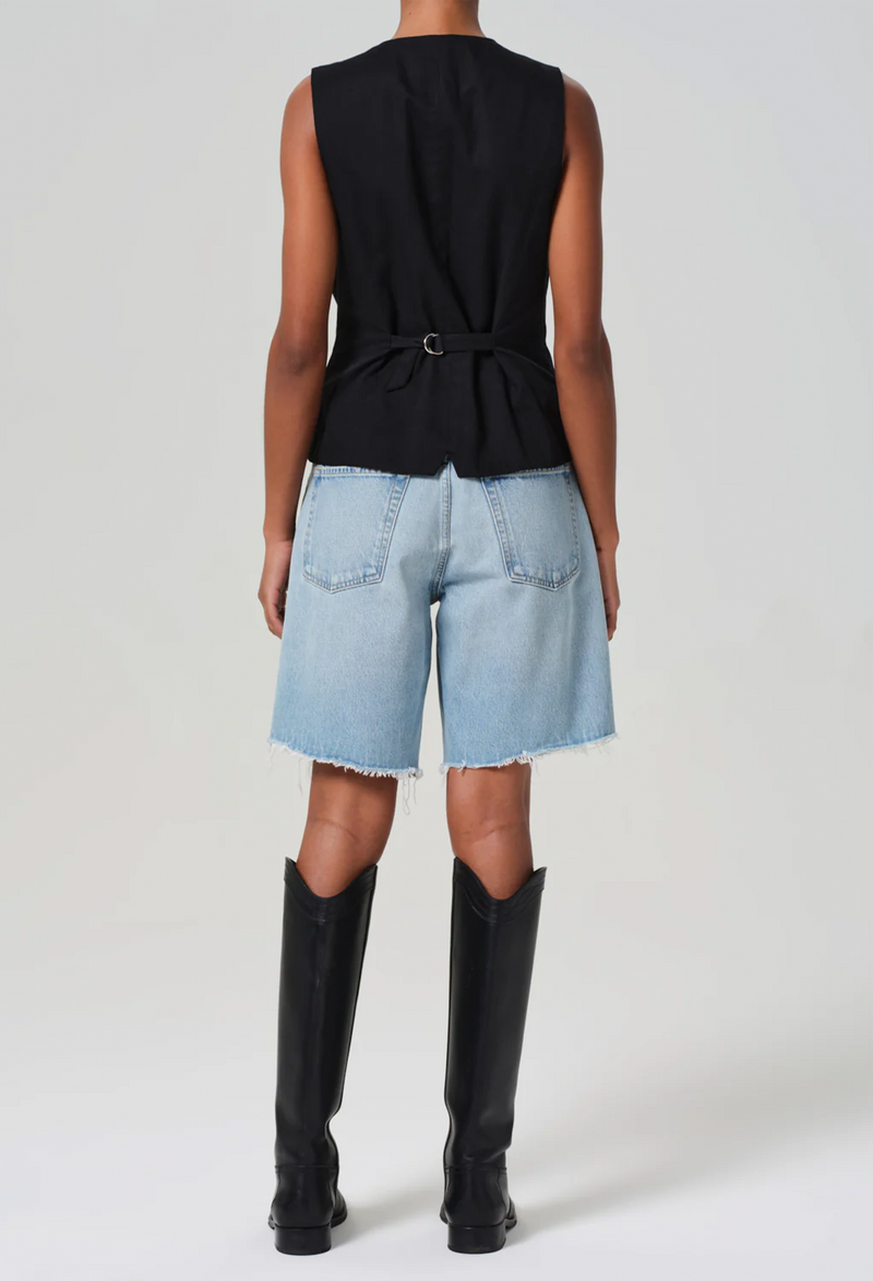 the back of a model wearing a black vest and long light wash denim shorts with a frayed hem. white background.