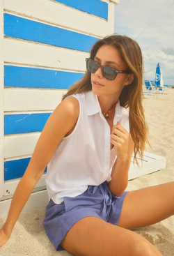 a model sitting on the beach wearing a white sleeveless button-front shirt with chest patch pocket and indigo shorts. 