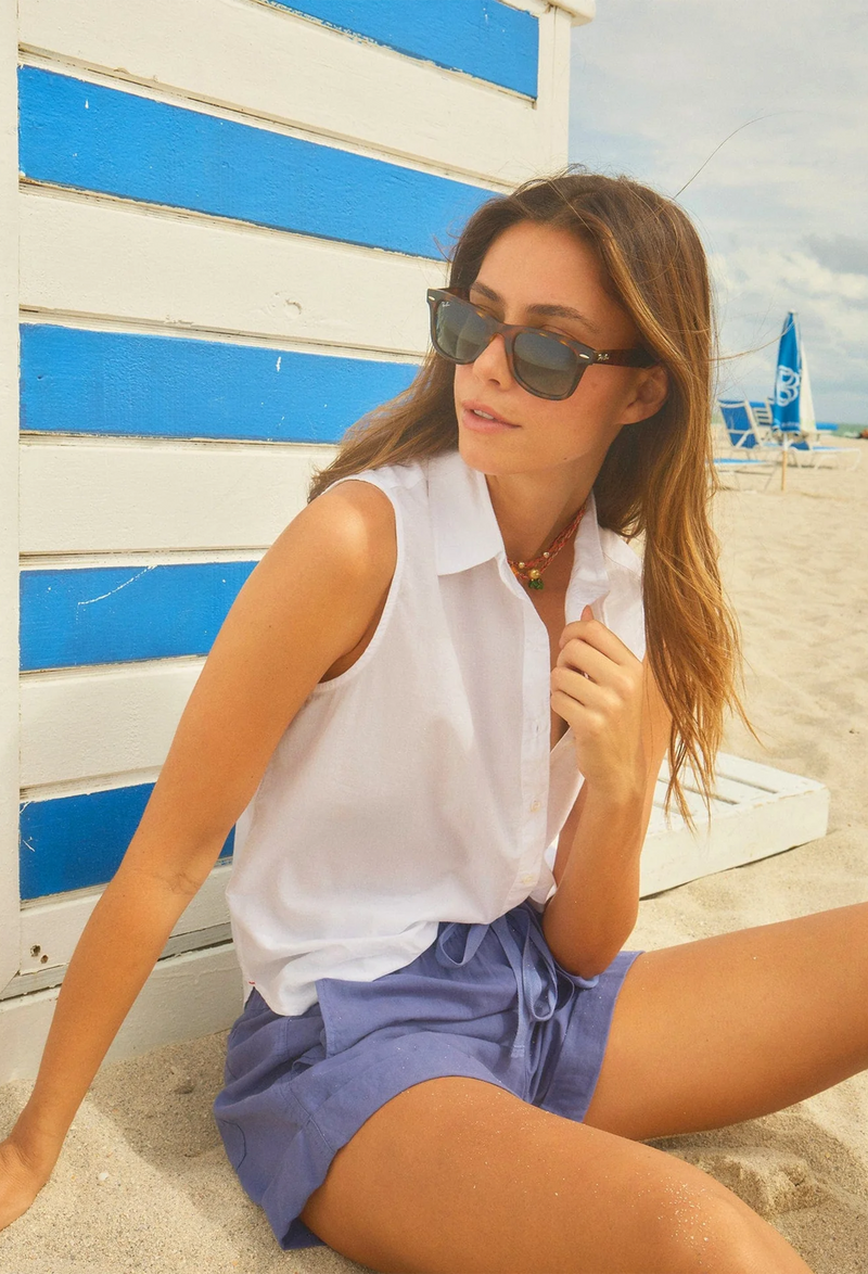 a model sitting on the beach wearing a white sleeveless button-front shirt with chest patch pocket and indigo shorts. 