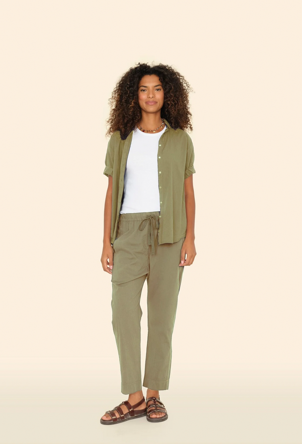 a model wearing a white top and open olive green button down shirt with matching olive green pull on drawstring pants. tan background.