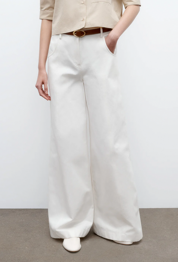 a model wearing wide leg white pants with a belt and beige top. white background.