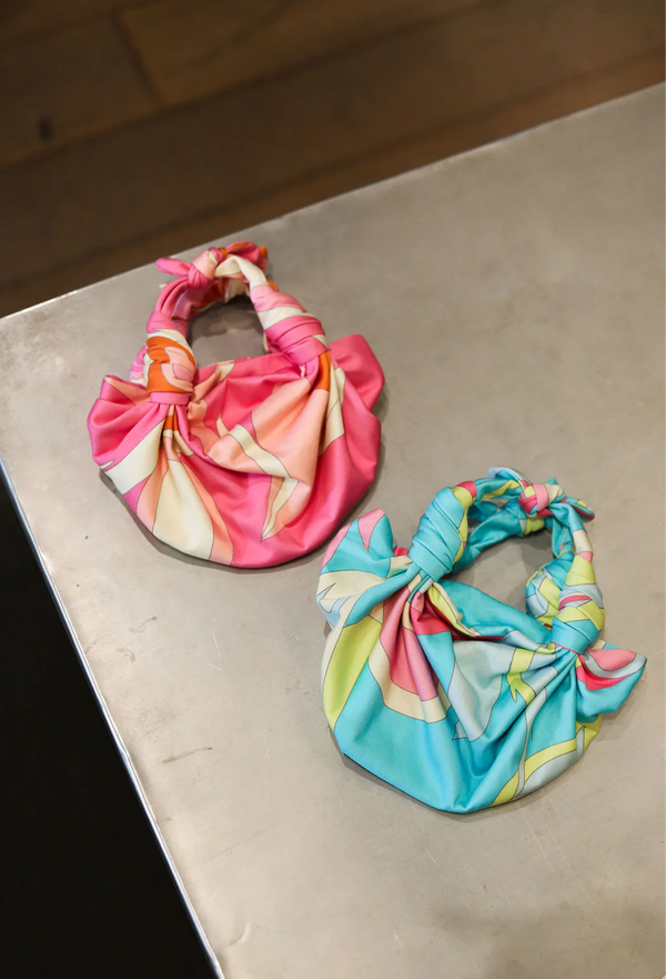 two bags on a tabletop. a pink knotted strap handbag with white and orange and a blue knotted strap handbag with pink and green. 
