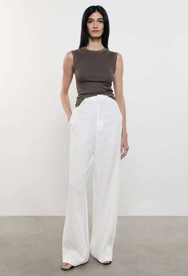 a model wearing a brown tank top and white linen pants. white background.