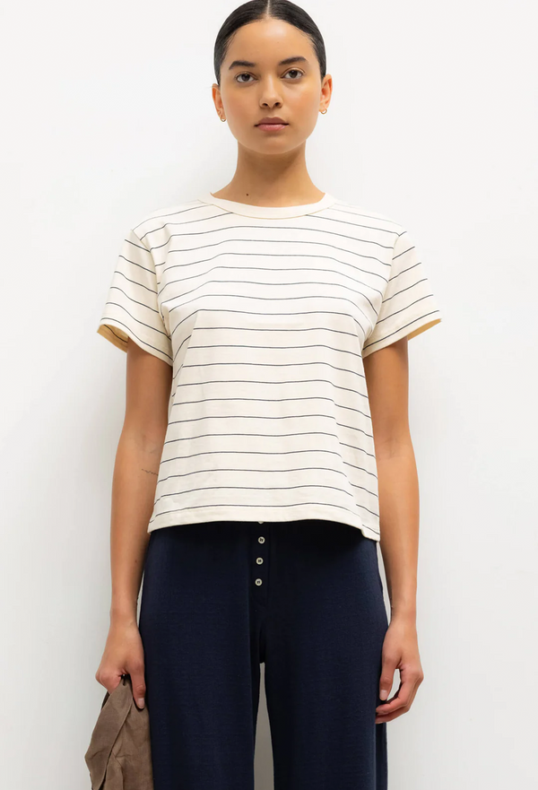 a model wearing an off-white short sleeve t-shirt with thin dark blue stripes and blue pants. white background.