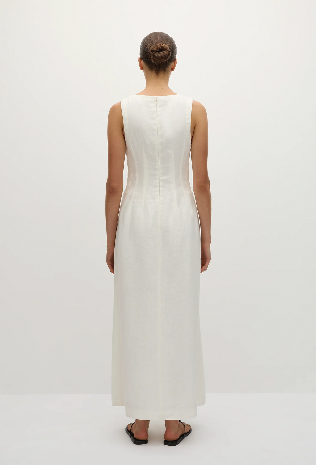 the back of a model wearing a sleeveless deep v-neck white maxi dress with a fitted bodice and slightly flared skirt. white background.