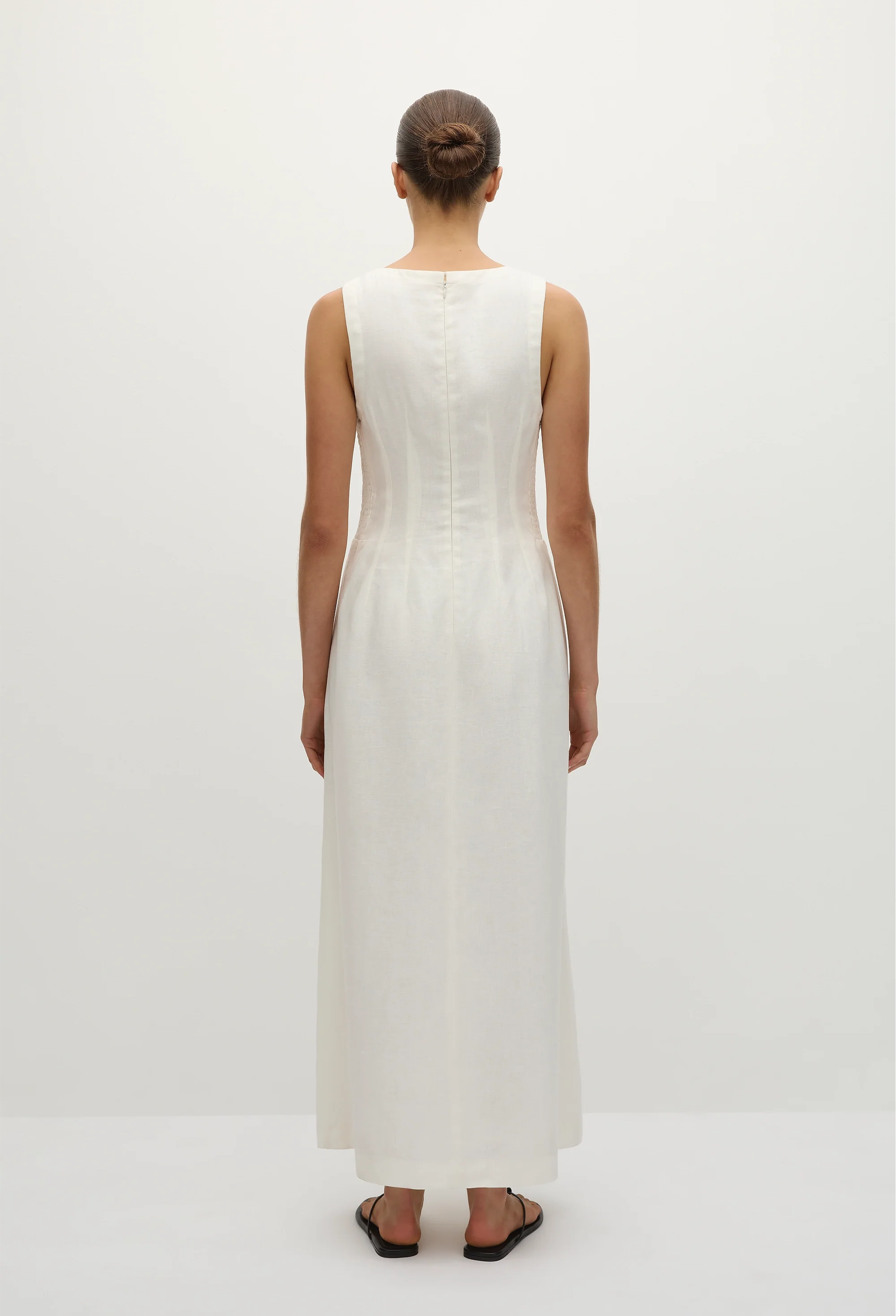 the back of a model wearing a sleeveless deep v-neck white maxi dress with a fitted bodice and slightly flared skirt. white background.