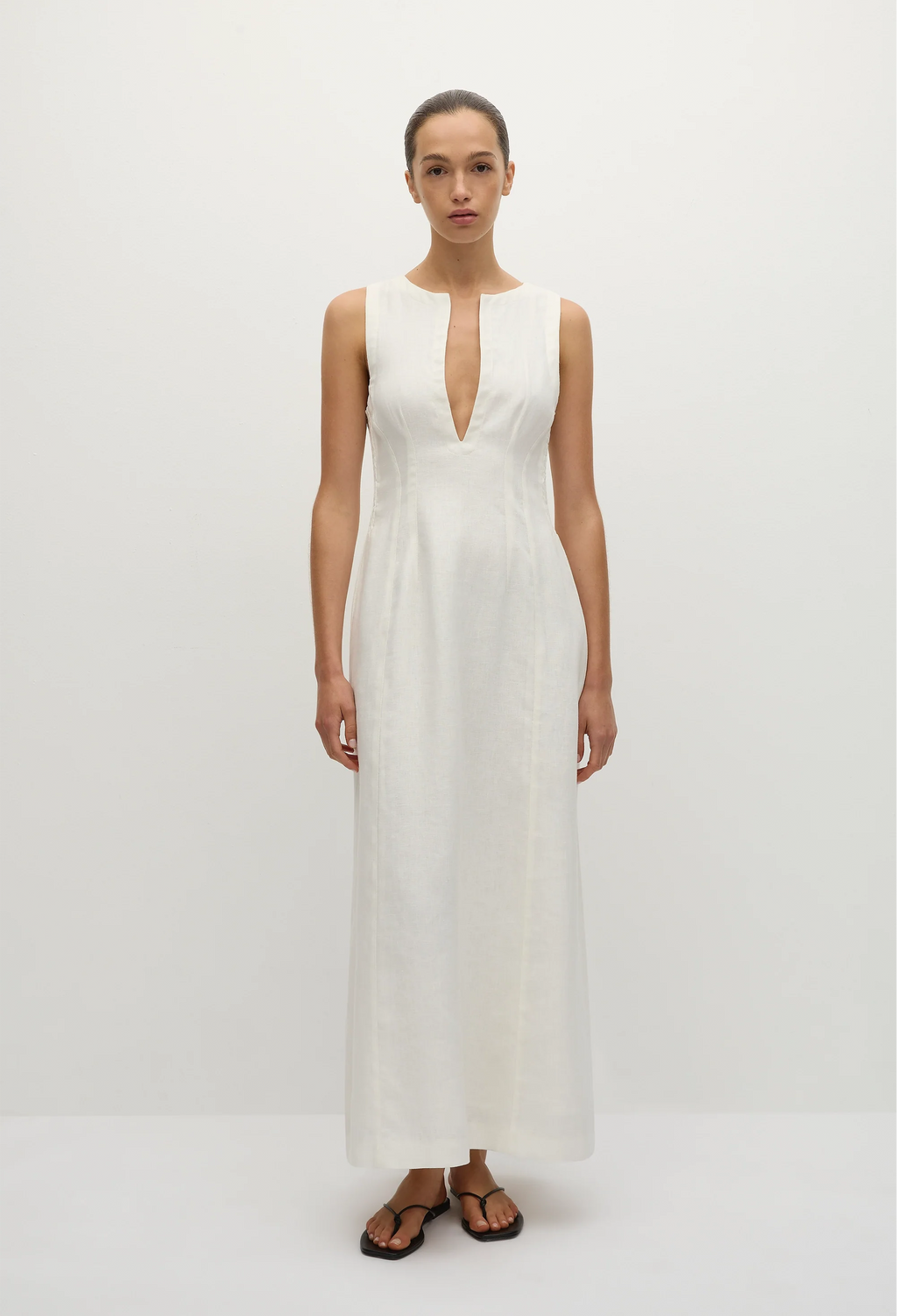 a model wearing a sleeveless deep v-neck white maxi dress with a fitted bodice and slightly flared skirt. white background.