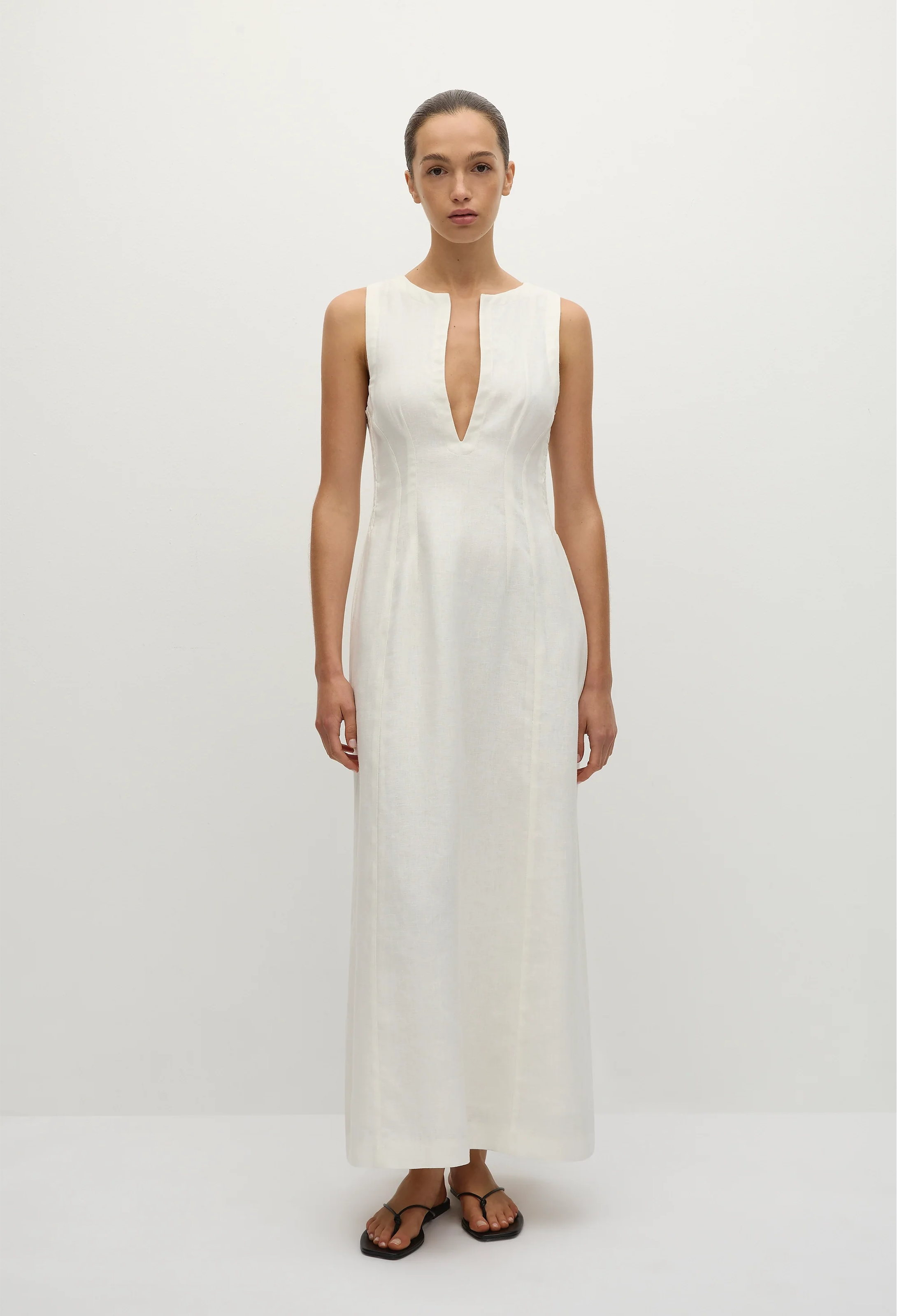 a model wearing a sleeveless deep v-neck white maxi dress with a fitted bodice and slightly flared skirt. white background.