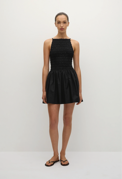 a model wearing a black mini dress with skinny straps and a shirred bodice with a flared a-line skirt. white background.