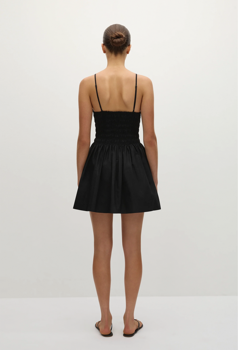 the back of a model wearing a black mini dress with skinny straps and a shirred bodice with a flared a-line skirt. white background.