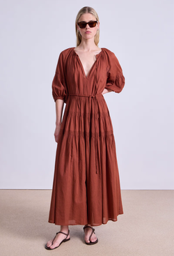 a model wearing a rust colored puff sleeve v-neck maxi dress with a tiered skirt and tie belt. white background.