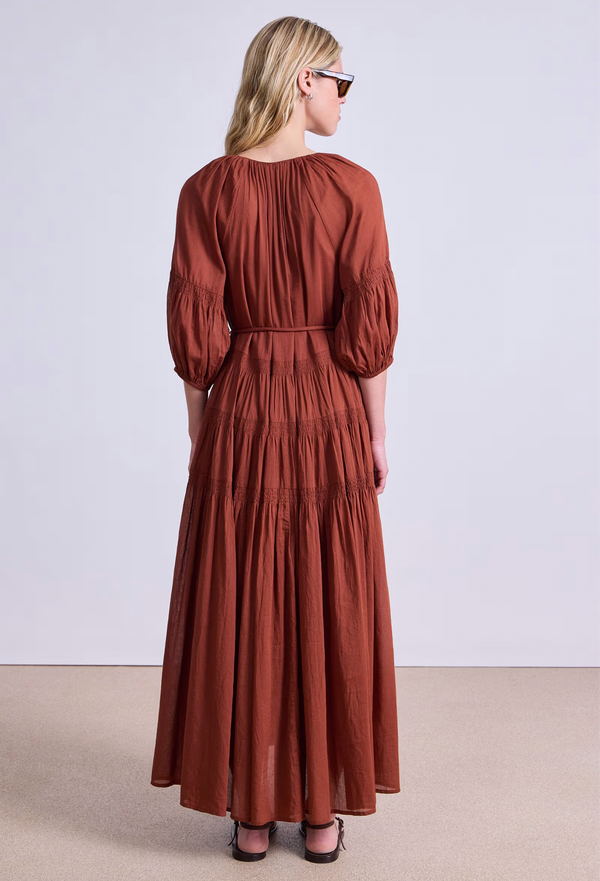the back of a model wearing a rust colored puff sleeve v-neck maxi dress with a tiered skirt and tie belt. white background.