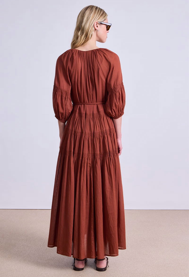 the back of a model wearing a rust colored puff sleeve v-neck maxi dress with a tiered skirt and tie belt. white background.