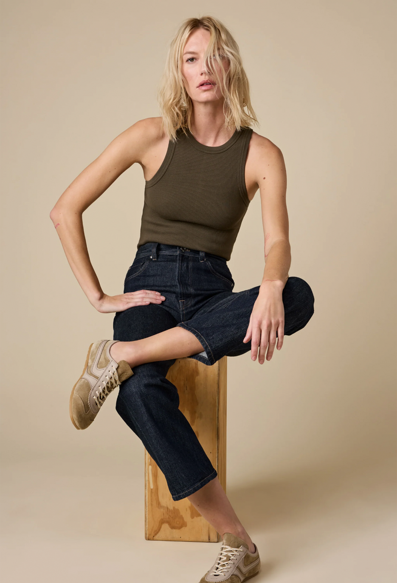 a model wearing an olive high neck racerback ribbed tank top and jeans. beige background