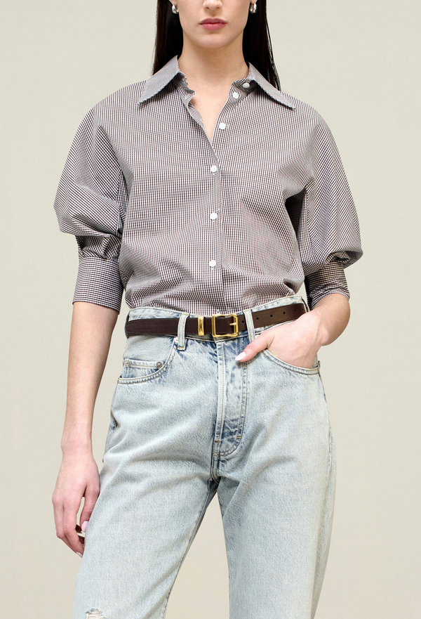 a model wearing a brown gingham 3/4 puff sleeve button down top with jeans. tan background.