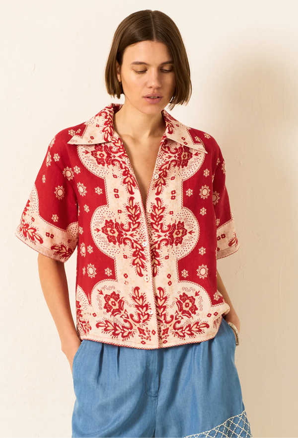 a model wearing a button up red bandana collared shirt with blue bottoms. white background.
