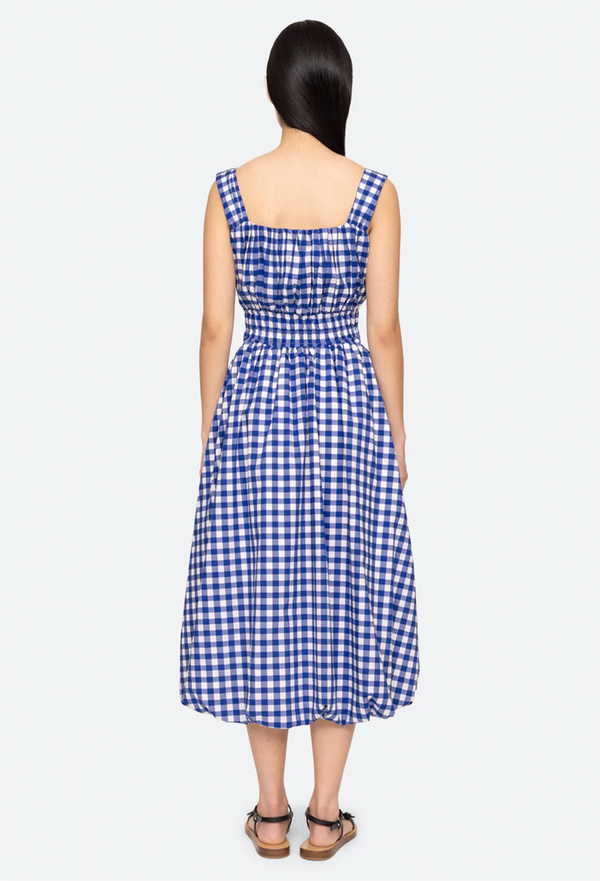 the back of a model wearing a blue gingham midi dress with thick straps and a cinched waist with a bubble hem. white background.