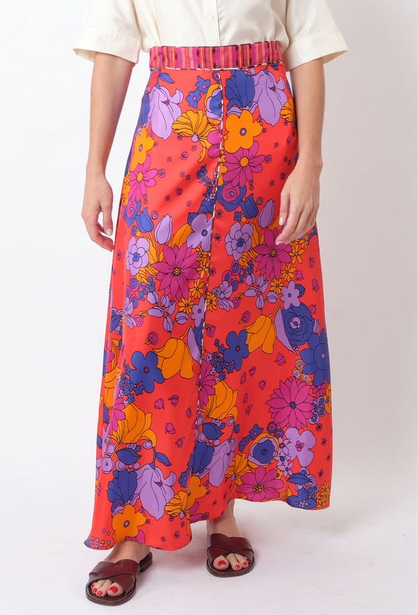 a model wearing a button down shirt and a long red skirt with multicolored floral print. white background.