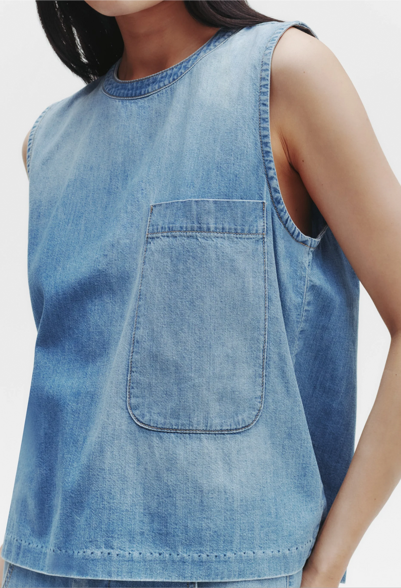 a model wearing a sleeveless denim shirt with a rounded neckline and a deep chest patch pocket. white background