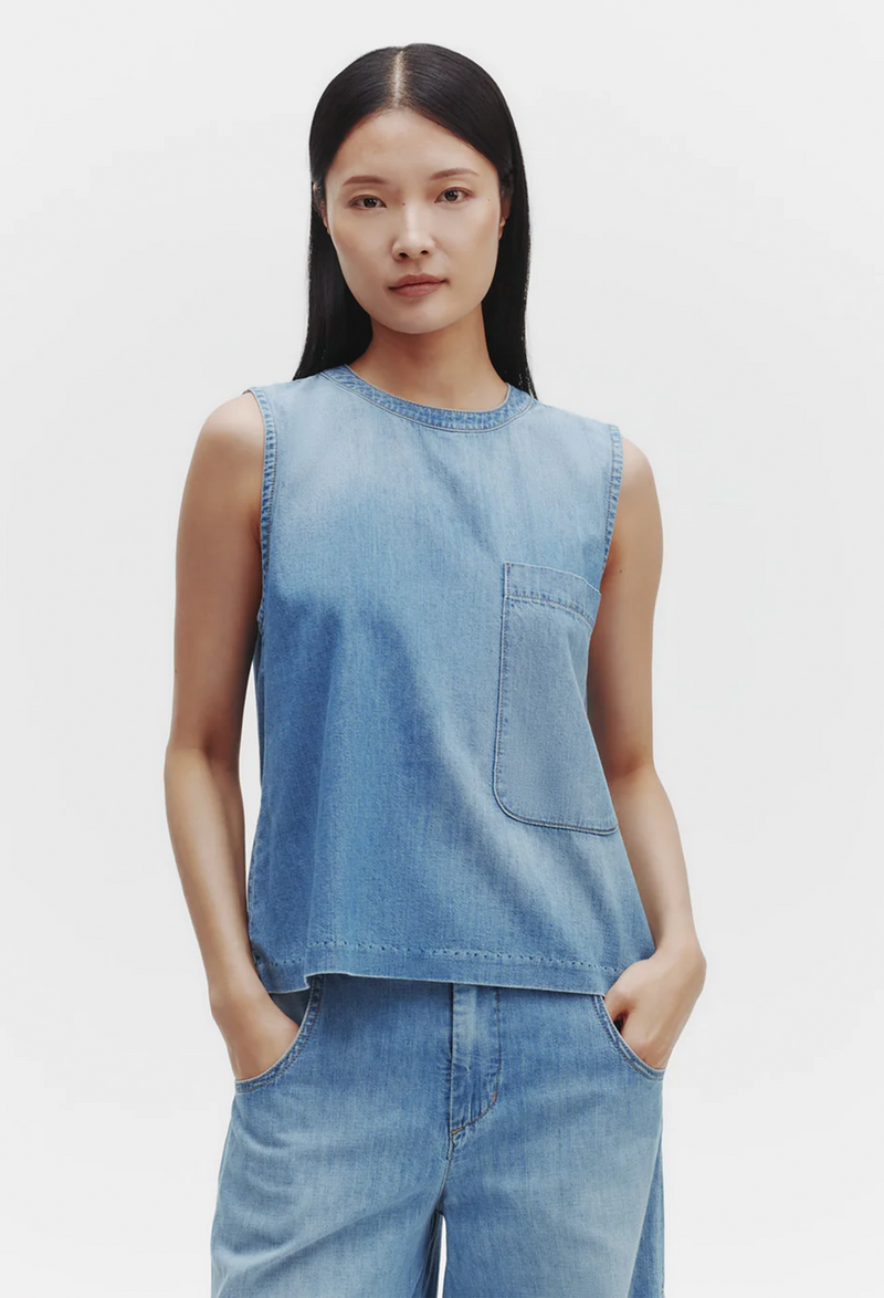 a model wearing a sleeveless denim shirt with a rounded neckline and a deep chest patch pocket. white background