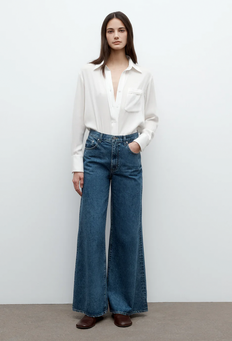 a model wearing a button down ivory collared silk shirt with a chest patch pocket and jeans. white background