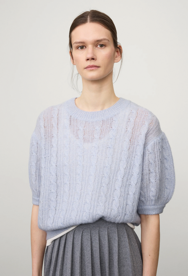Woman wearing a light blue cable knit 3/4 sleeve sweater with gray pleated skirt on a white background