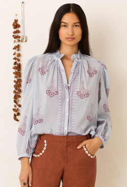 Woman wearing a light blue blouse with floral embroidery and brown pants against a beige background