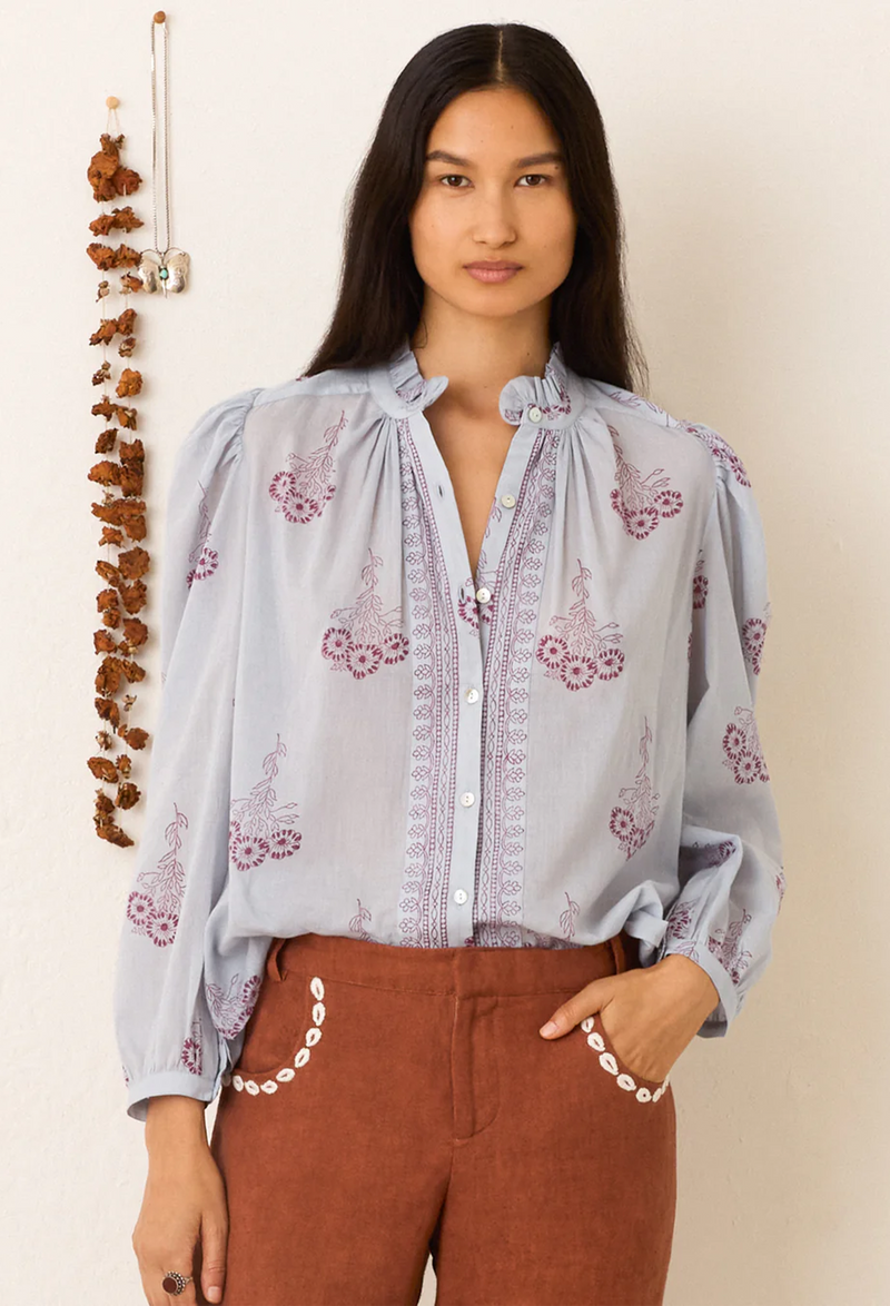 Woman wearing a light blue blouse with floral embroidery and brown pants against a beige background