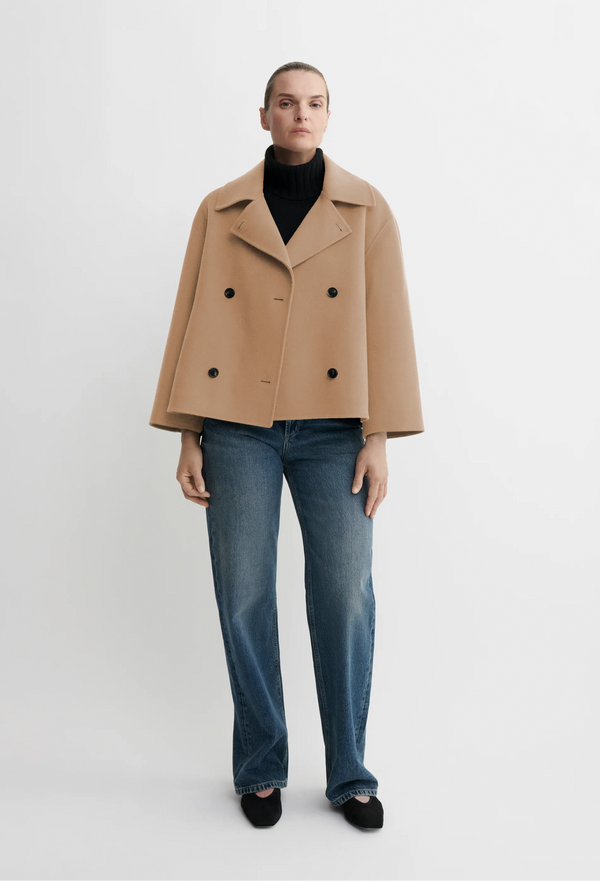 Person wearing a beige coat, black turtleneck, and blue jeans on a white background