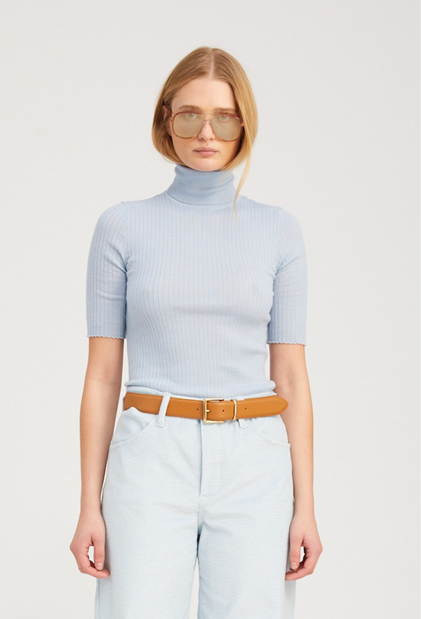 Woman wearing a light blue ribbed turtleneck sweater and light blue pants with a brown belt on a white background.