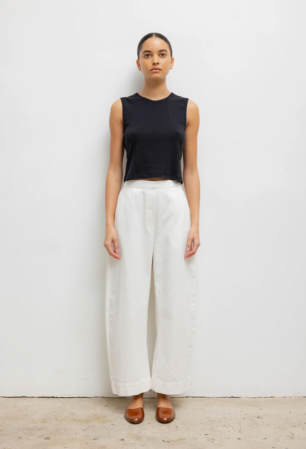 Woman wearing a black sleeveless top and white barrel leg pants on a plain background
