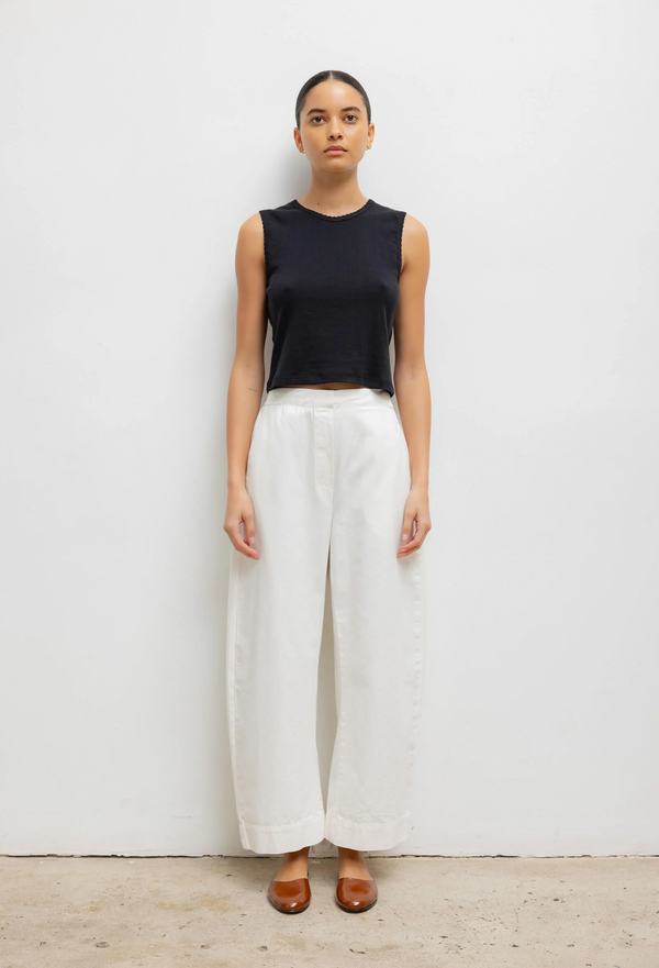 Woman wearing a black sleeveless top and white barrel leg pants on a plain background