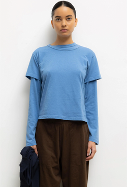 Person wearing a blue long-sleeve shirt under a blue short sleeve tee and brown pants against a white background