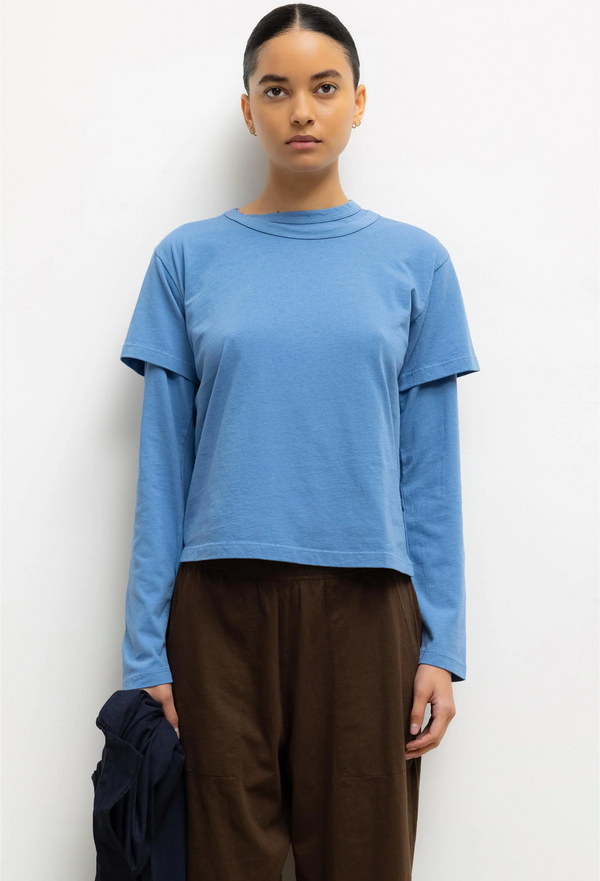 Person wearing a blue long-sleeve shirt under a blue short sleeve tee and brown pants against a white background