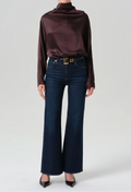 A model wearing a dark plum satin long-sleeve turtleneck top, dark blue wide-leg denim jeans with a gold buckle belt, and black pointed-toe heels.