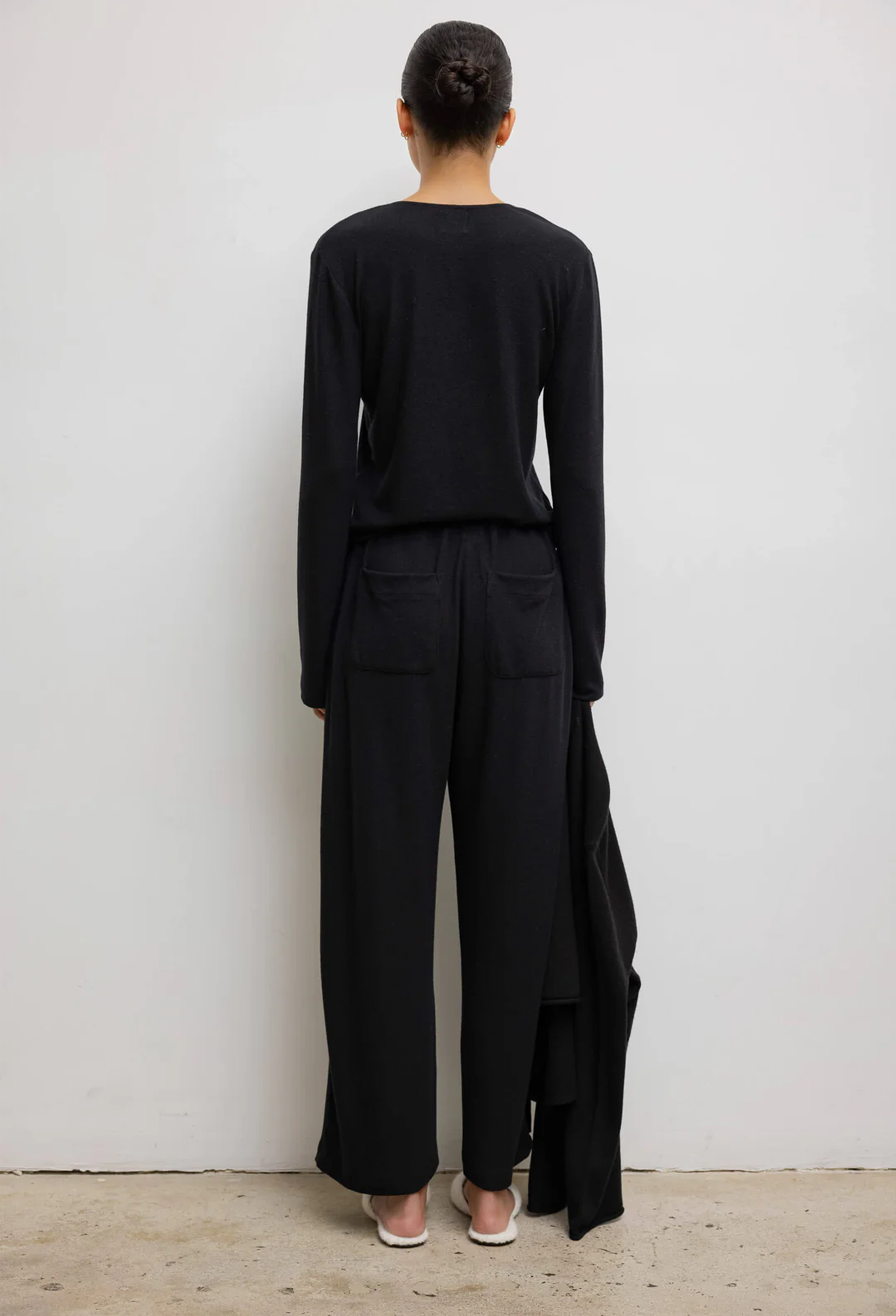 From backside model wears black long sleeve top. She also wears black drawstring pants with back pockets and white shoes. She stands in front of a white wall. 
