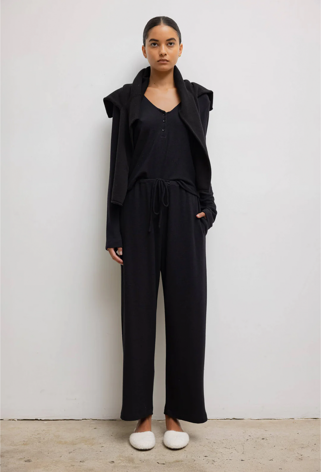 Model wears black long sleeve top with three front buttons with black sweater around her shoulders. She also wears black drawstring pants and white shoes. She stands against a white wall. 