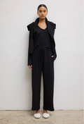 Model wears black long sleeve top with three front buttons with black sweater around her shoulders. She also wears black drawstring pants and white shoes. She stands against a white wall. 