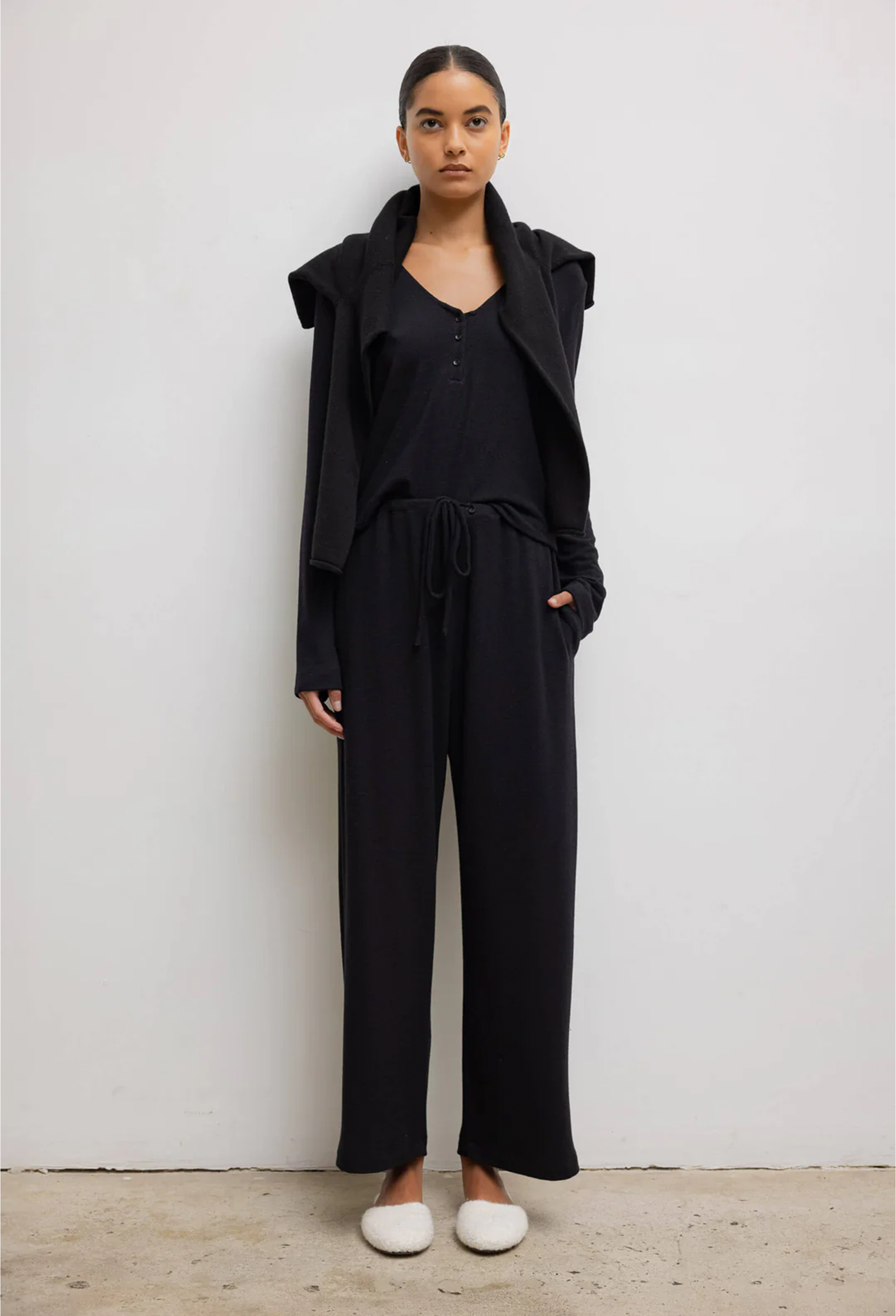 Model wears black long sleeve top with three front buttons with black sweater around her shoulders. She also wears black drawstring pants and white shoes. She stands against a white wall. 