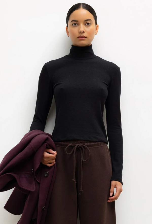 Person wearing a black turtleneck and brown pants holding a purple coat against a white background