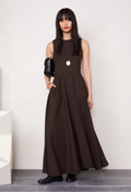 Model wears long brown sleeveless dress with black boots and holds a black purse while wearing a long necklace. She stands in front of a white wall. 