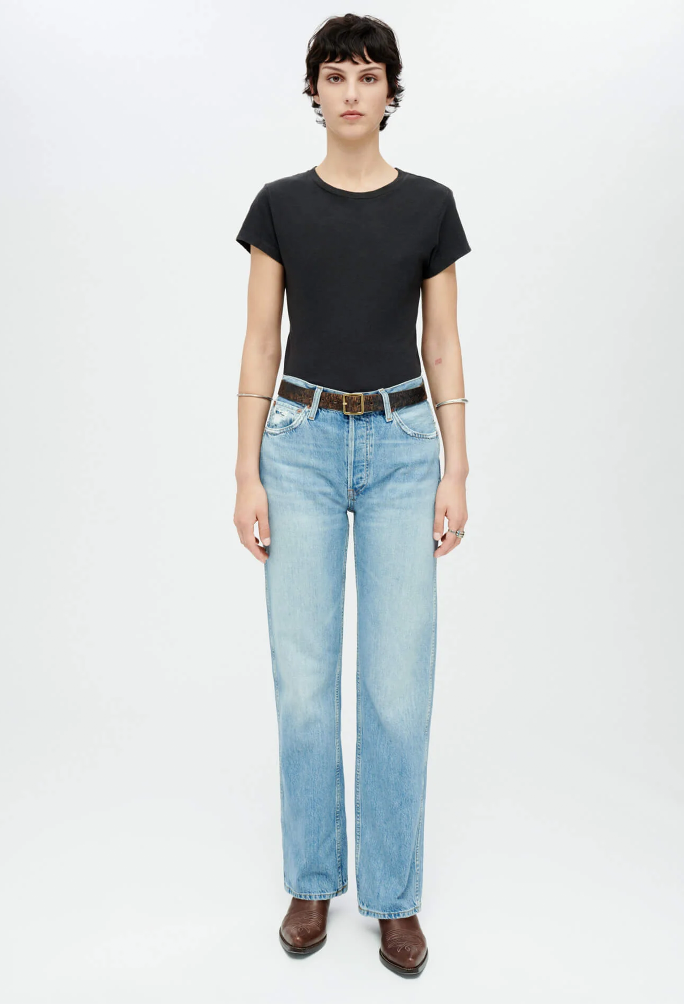 Model wears black short sleeved t-shirt with light wash jeans. She wears a silver bangle on each arm, brown boots, and a brown belt. Set against a white background.