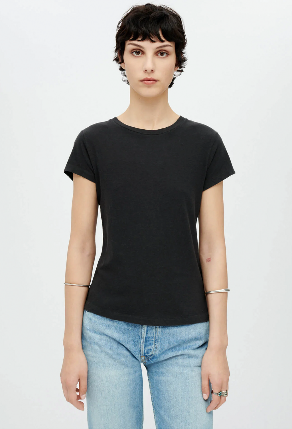 Model wears black short sleeved t-shirt with light wash jeans. She wears a silver bangle on each arm. Set against a white background.