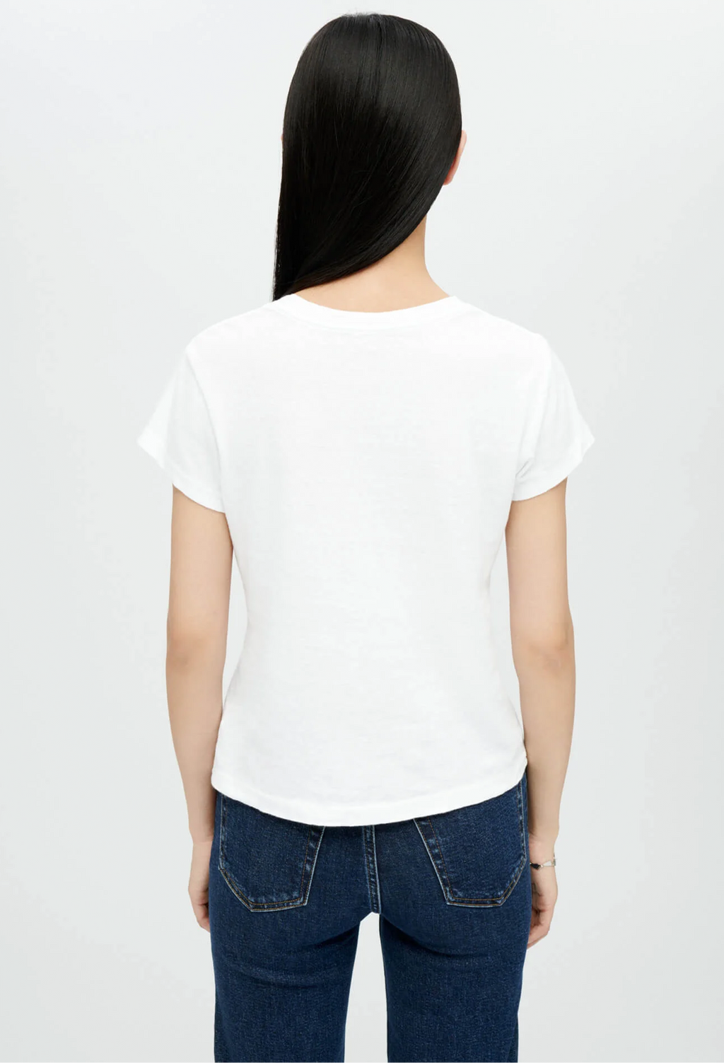 Backside of model wearing white crewneck t-shirt with dark wash blue jeans. She stands in front of a white background. 