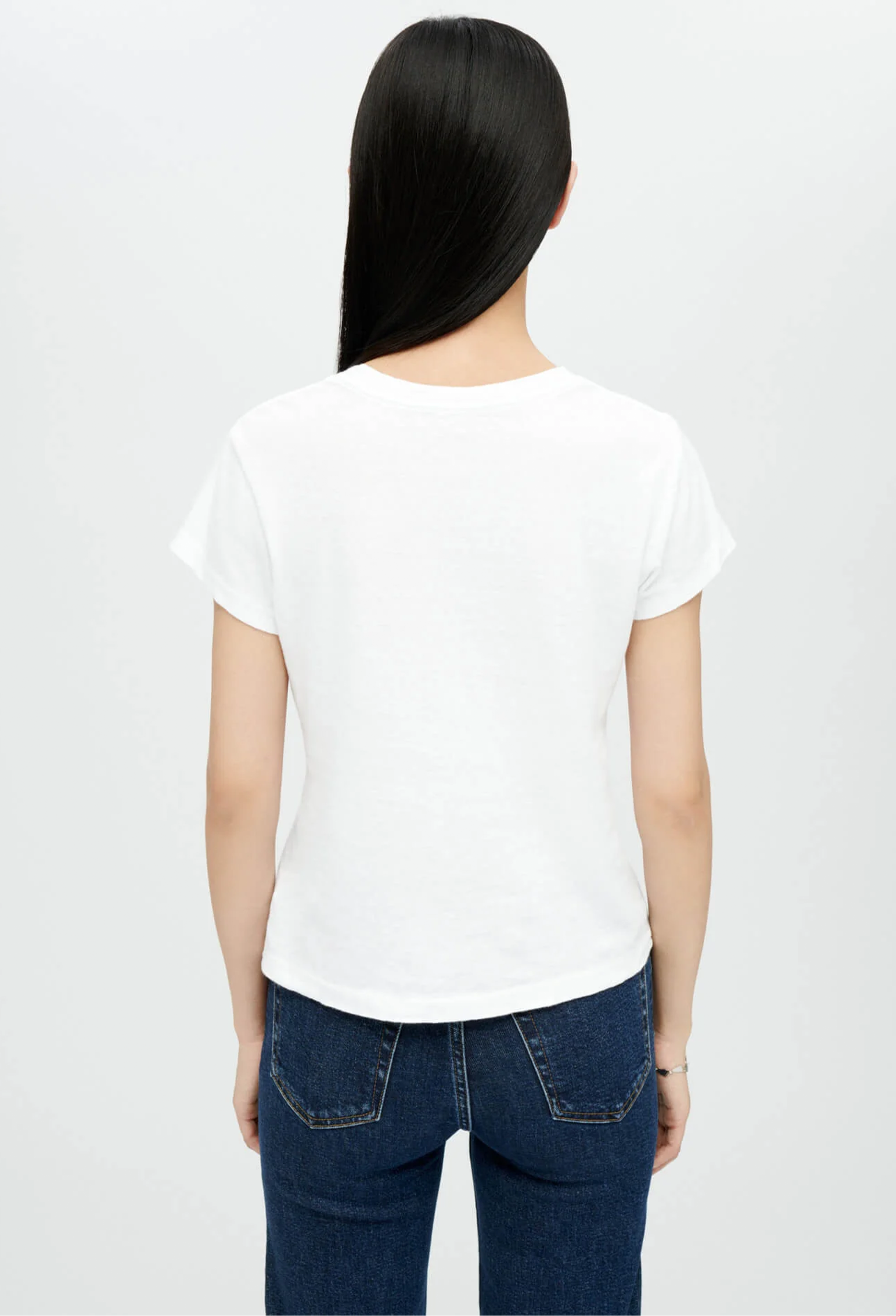 Backside of model wearing white crewneck t-shirt with dark wash blue jeans. She stands in front of a white background. 