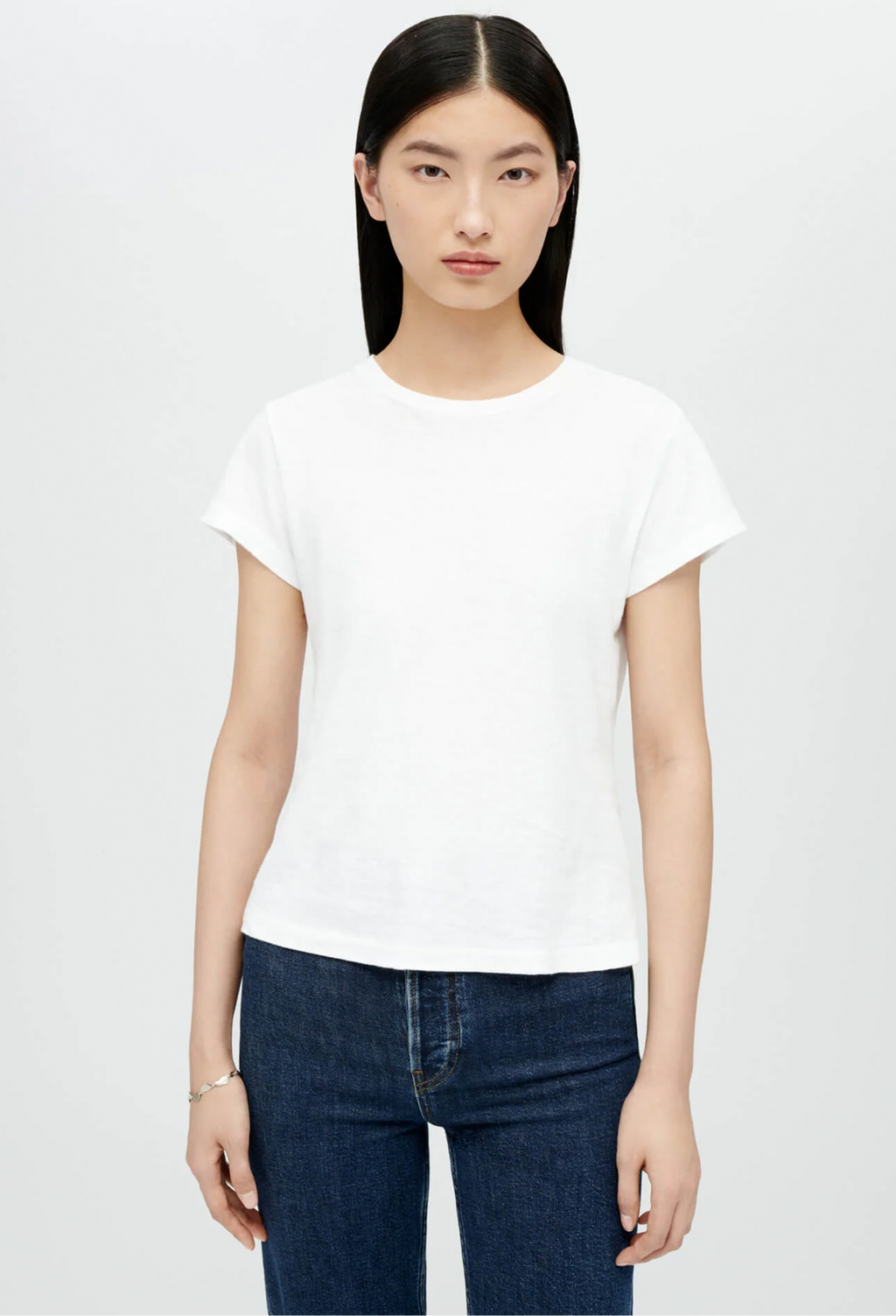 Model wears white crewneck t-shirt with dark wash blue jeans. She stands in front of a white background. 
